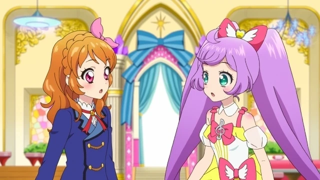 Escena de Akari Ozora y Laala Manaka dialogando con expresiones de asombro en un elegante salón, simbolizando el cruce inesperado de sus mundos idol en Aikatsu! × PriPara The Movie.