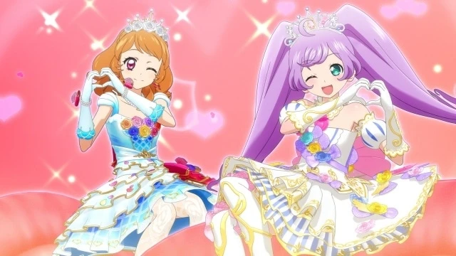 Ilustración promocional de Aikatsu! × PriPara The Movie con Akari Ozora y Laala Manaka posando con trajes brillantes y coronas, formando un corazón con las manos sobre un fondo rosa lleno de destellos, simbolizando la unión entre ambas franquicias.